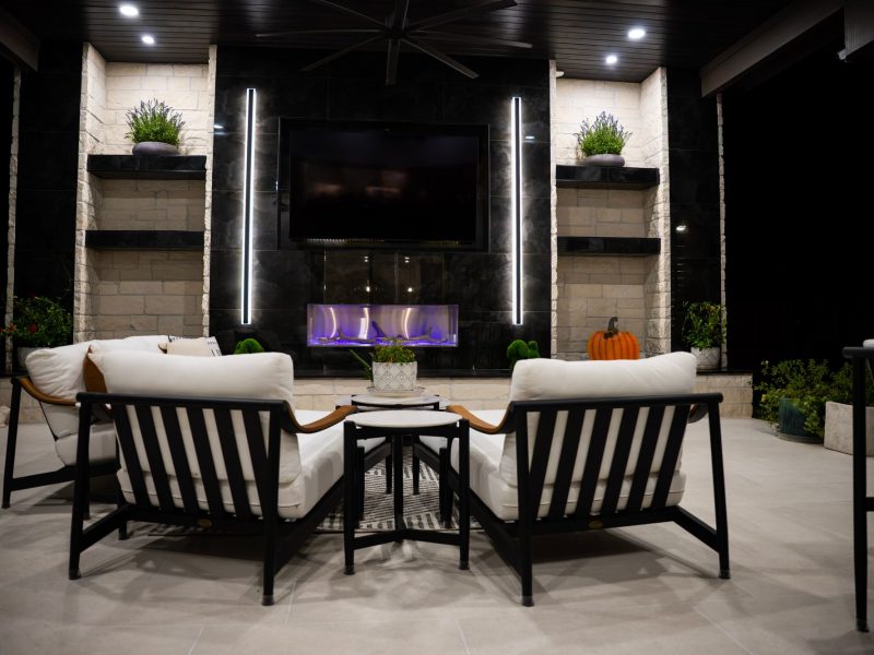 Modern outdoor patio with a stone wall, linear fireplace, black armchairs with white cushions, and a small side table.