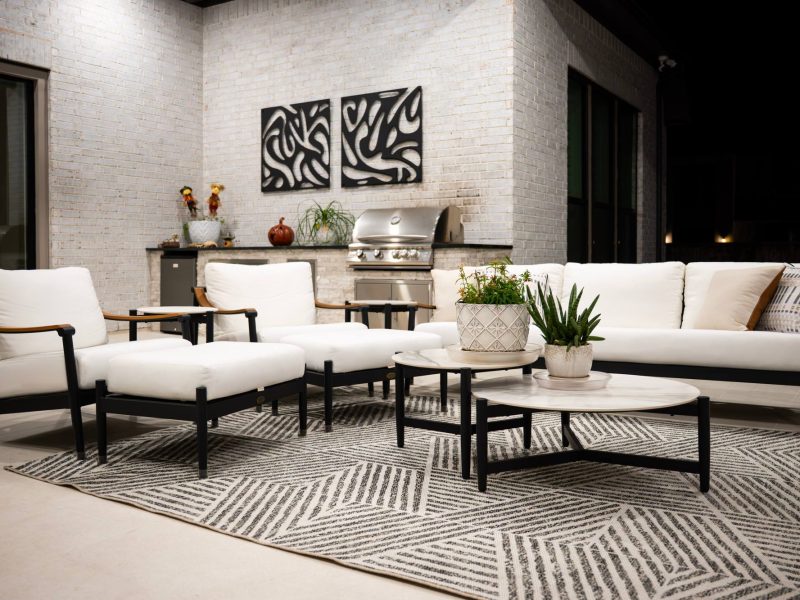 Modern outdoor patio with white cushioned furniture, black frames, and a patterned rug against a white brick wall.