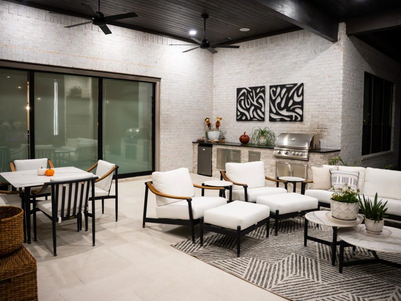 Modern covered patio with a dining set, white lounge seating, and an outdoor kitchen against a white brick wall.
