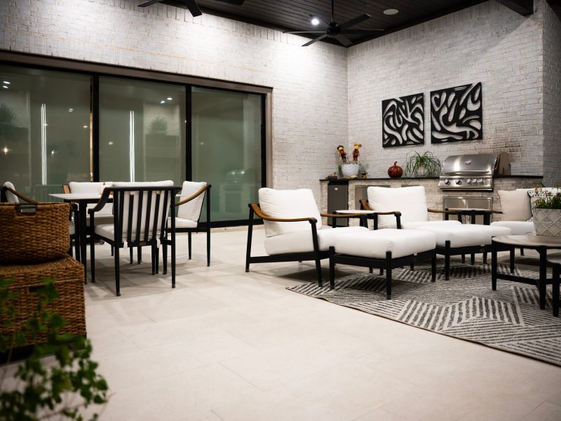 Covered patio with white modern lounge chairs, dining set, and built-in grill against white brick walls.