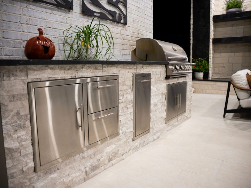 Outdoor kitchen with built-in grill and stainless steel storage cabinets against a white stone wall.