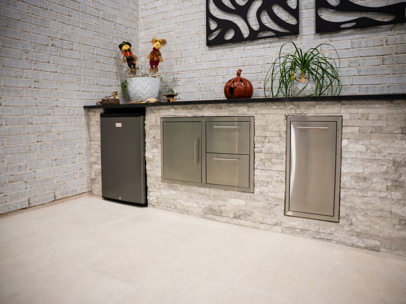 Outdoor kitchen with stainless steel appliances and a black countertop against a white brick wall.