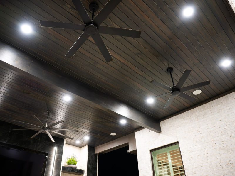 Stained wood patio ceiling with three modern black ceiling fans and recessed lighting.
