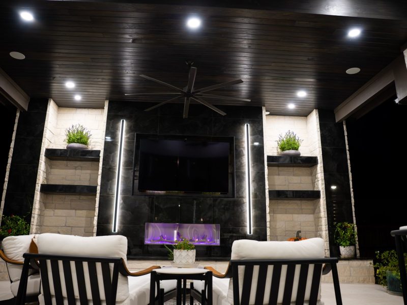 Modern outdoor patio with a black accent wall, built-in fireplace, large TV, and white lounge chairs.
