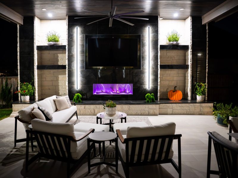 Modern covered patio with a linear purple fireplace, wall-mounted TV, and white lounge seating.