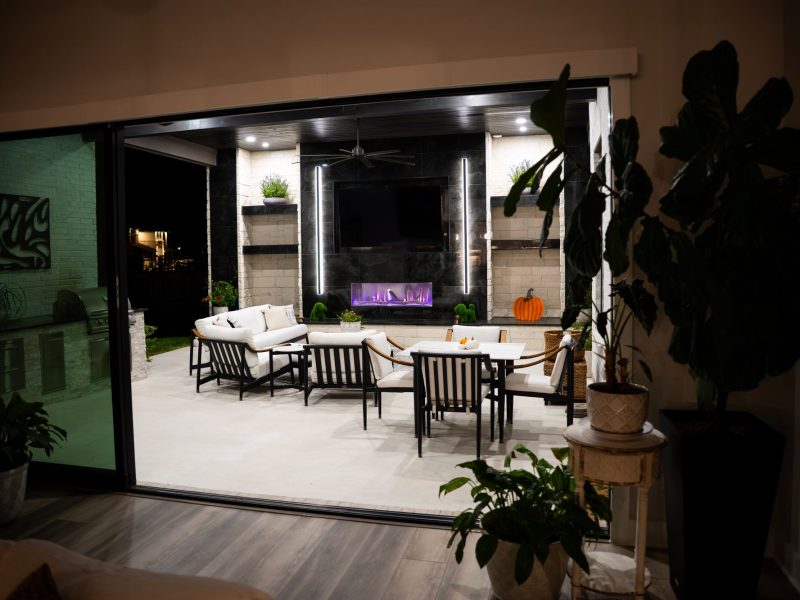 View of a covered patio with a modern fireplace, white lounge seating, and a dining set from inside the home.