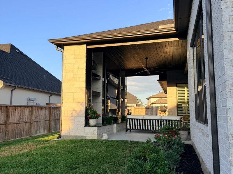 Covered outdoor patio with a modern fireplace, ceiling fan, and landscaping next to a white brick home.