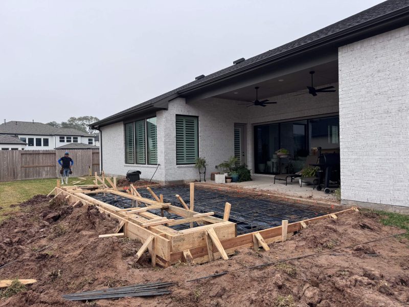Wooden forms and rebar framing for a new patio and fireplace extension against a white brick house.