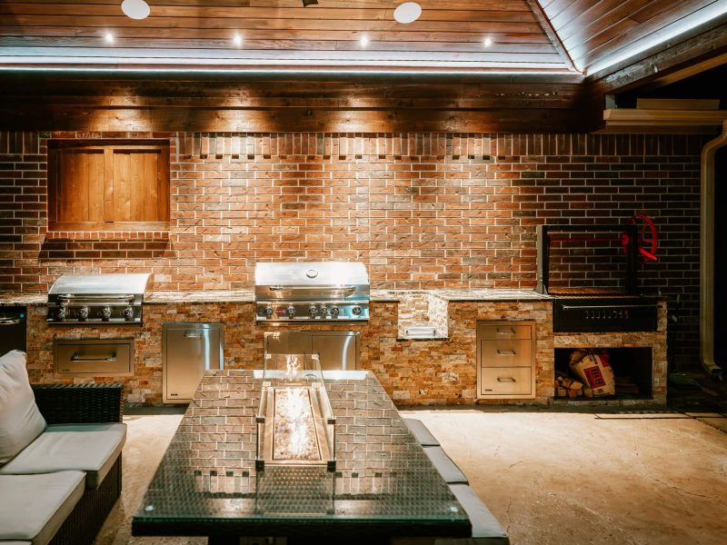 Outdoor kitchen with built-in grills and a fire pit table under a wood pavilion ceiling.