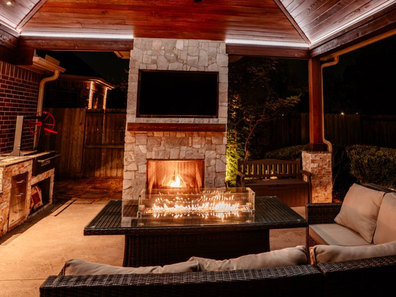Outdoor living area with a stone fireplace, mounted TV, and fire pit table under a wood pavilion.