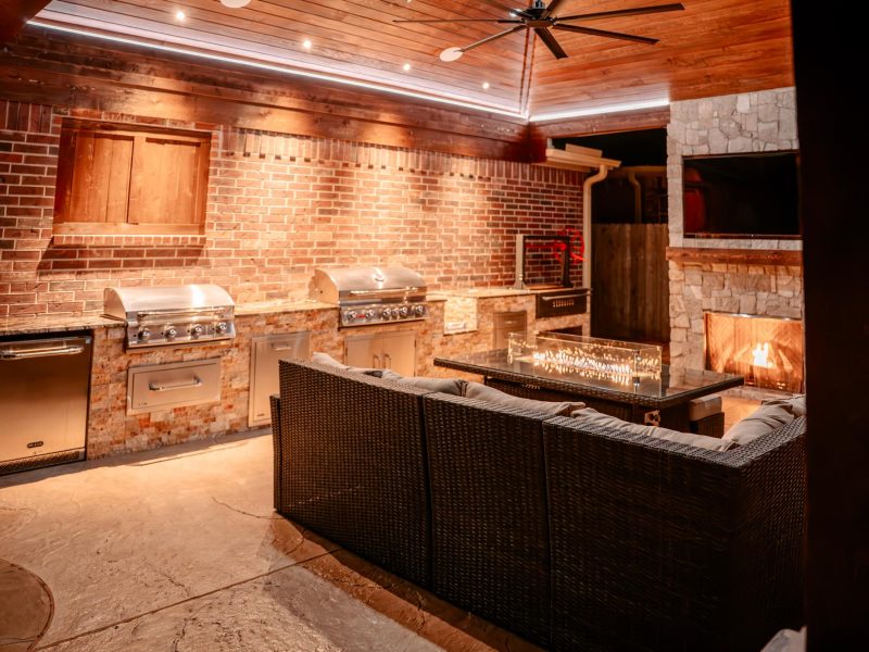 Outdoor kitchen with brick walls, dual grills, a wicker sofa, and a fireplace under a wood pavilion.