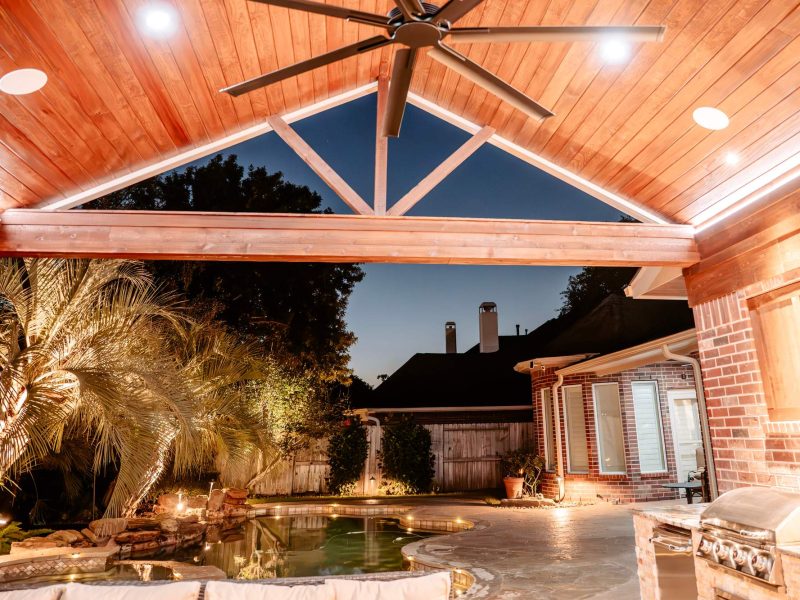 Vaulted wood pavilion ceiling with a large fan and recessed lights overlooking a swimming pool and outdoor kitchen.