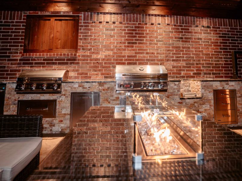Outdoor kitchen with brick walls, built-in grills, and a linear fire pit under a pavilion.
