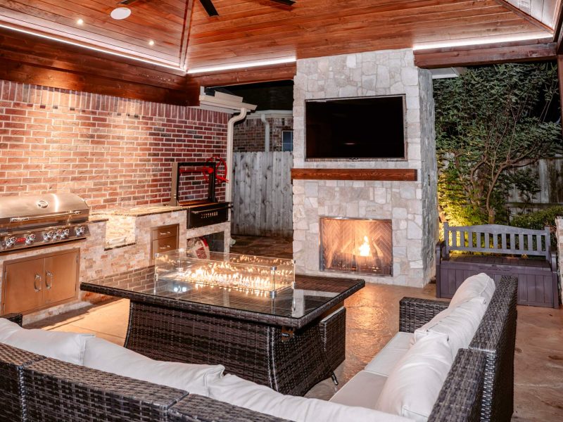 Outdoor kitchen and living area with a built-in grill, fire pit table, and stone fireplace with TV under a wood pavilion.