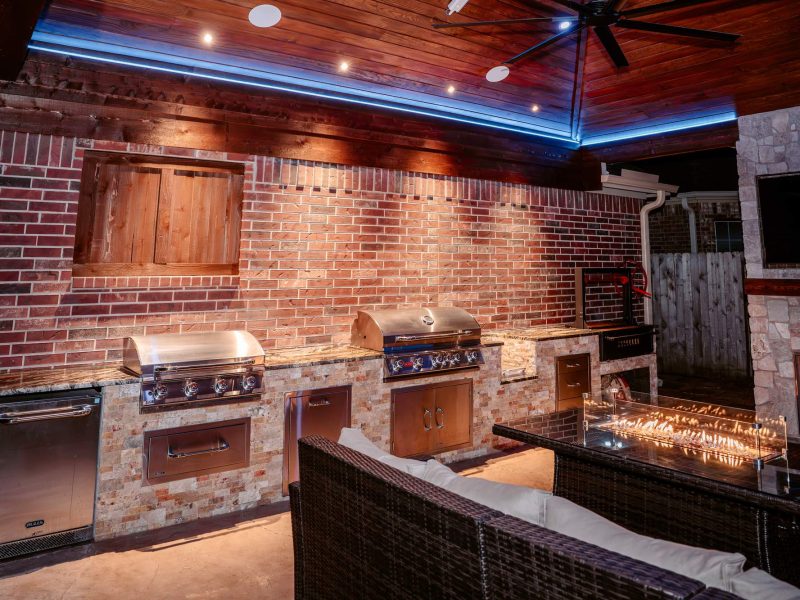 Outdoor kitchen with dual grills, stainless appliances, and a linear fire pit under a wood pavilion ceiling.