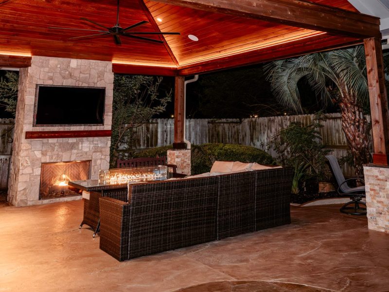 Outdoor living space with a stone fireplace, wall-mounted TV, and wicker sectional sofa under a wood pavilion.