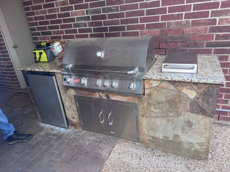 Outdoor kitchen with a built-in grill, small refrigerator, and granite countertop against a brick wall.