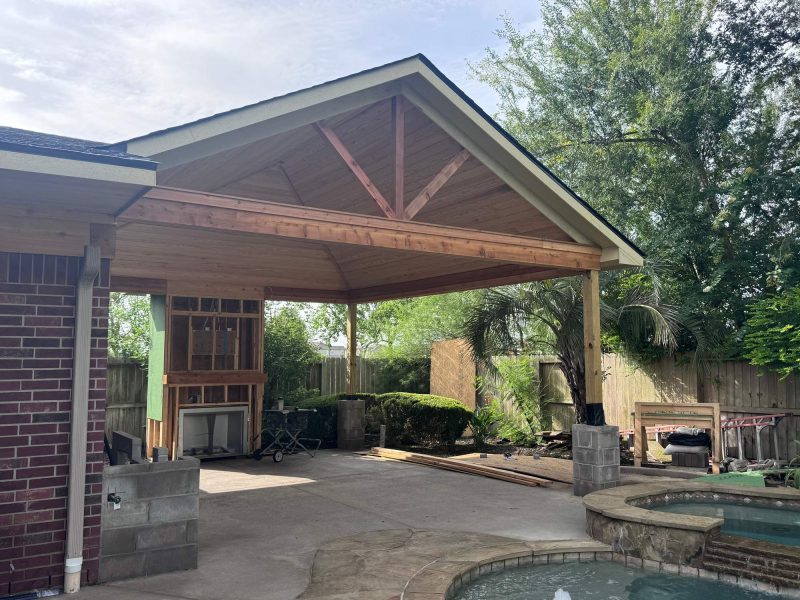 Unfinished wood pavilion structure with a vaulted ceiling over an outdoor patio and fireplace under construction.